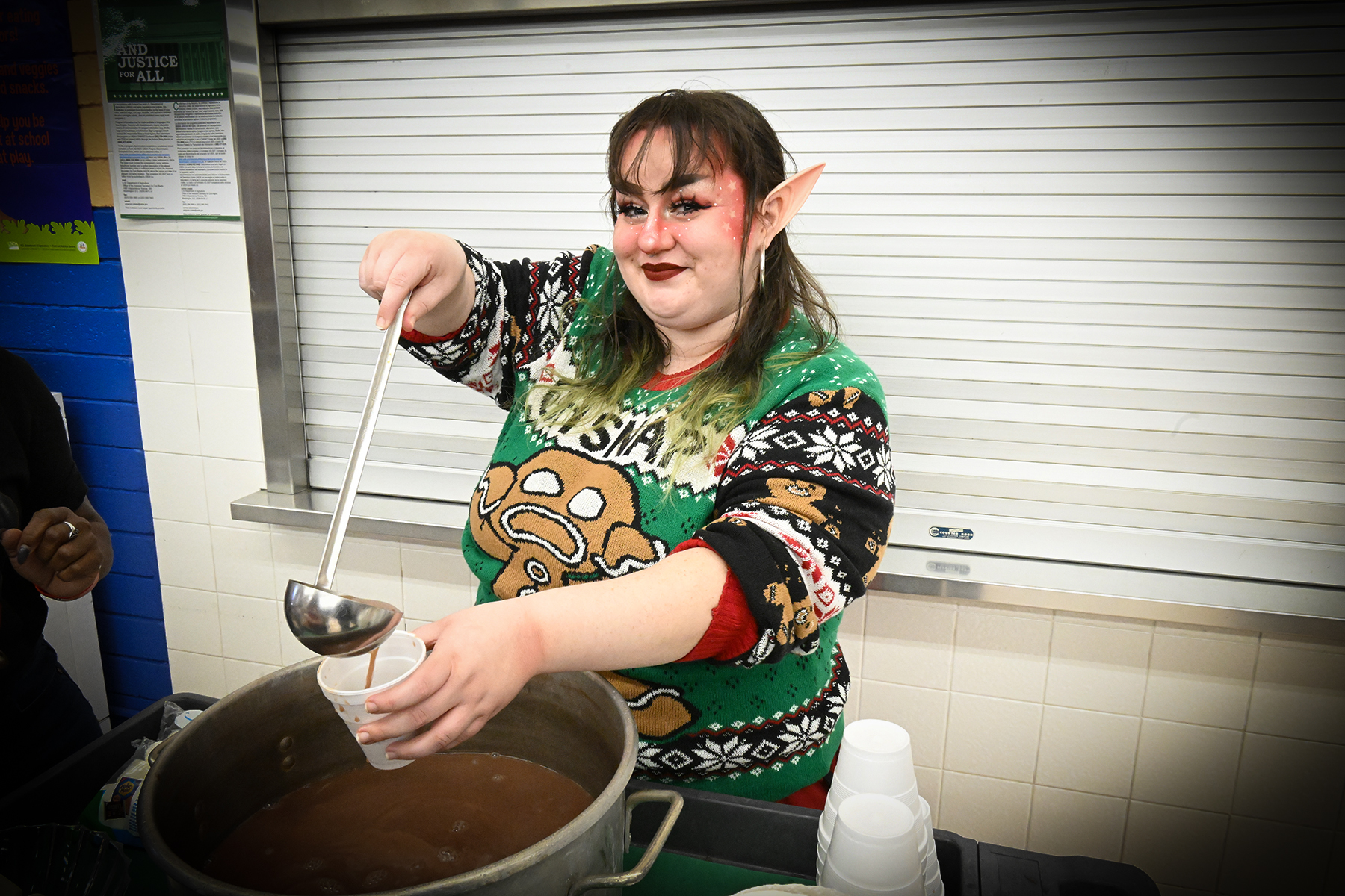 A woman wearing elf ears and a Christmas sweater serves up hot chocolate with a ladle