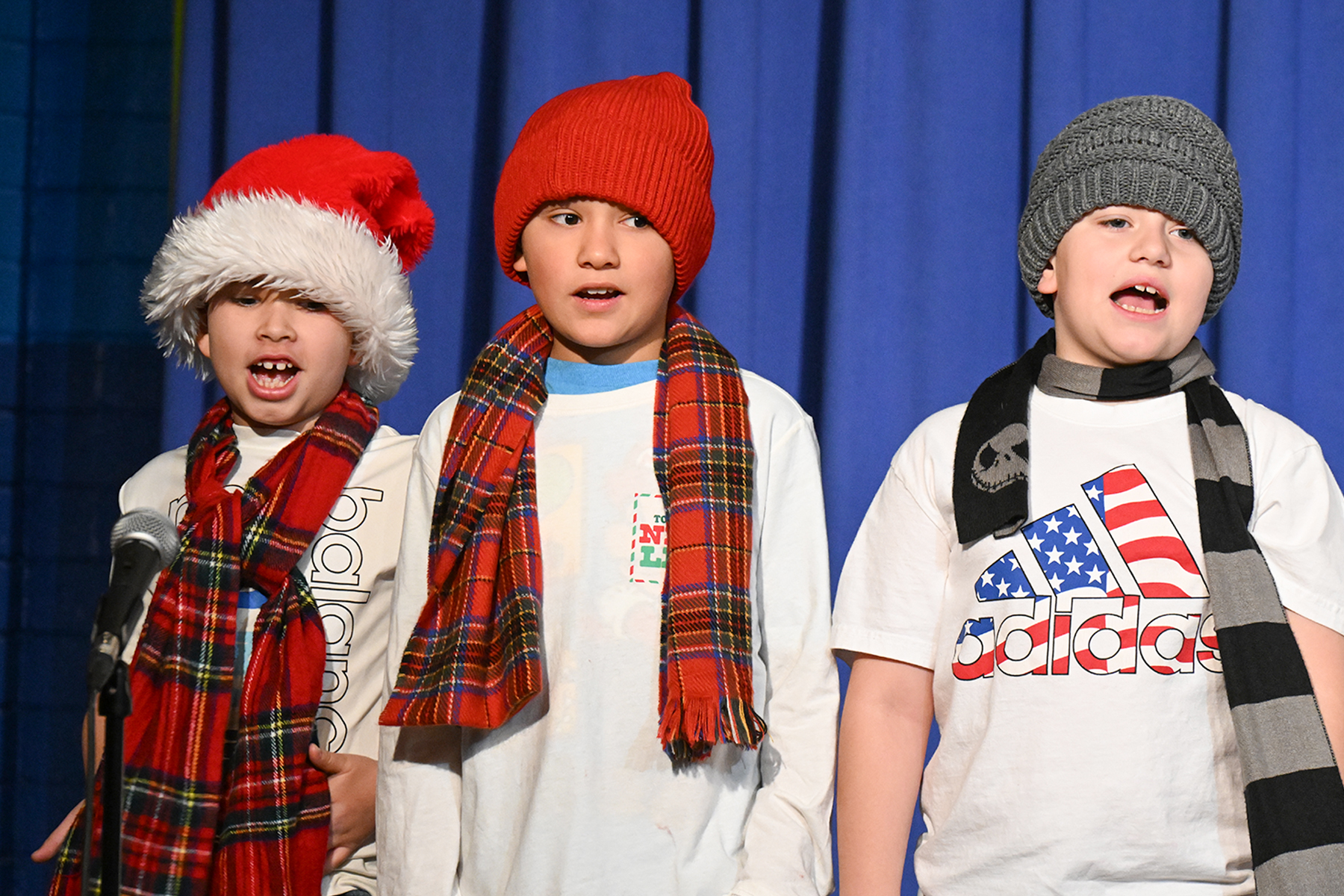 Three boys wearing winter hats and scarves sing on stage