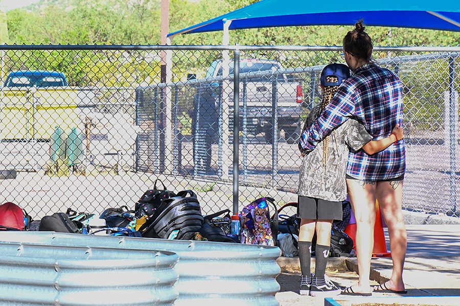 A boy and his mom hug while facing the fence lined with backpacks