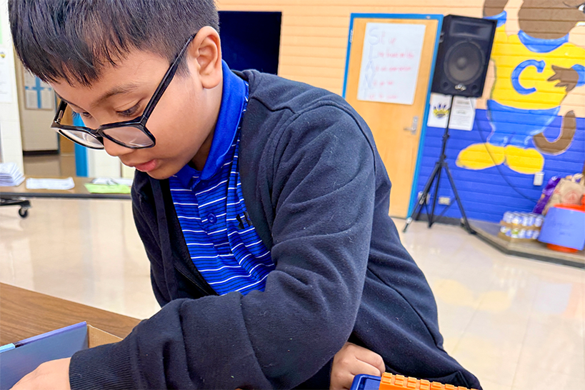 A boy with glasses and a blue striped shirt reaches into a box