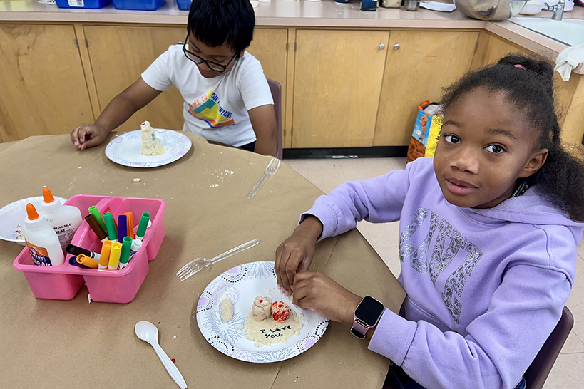 A girl in a purple hoodie looks up as she makes a masa sculpture at a table