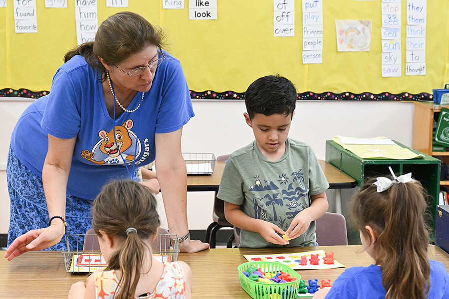 A woman in a blue shirt and skirt leans over the table to observe as kids work on a craft