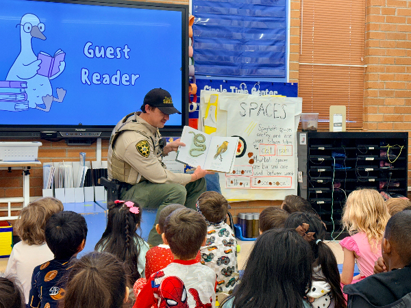 A man in a sheriff's uniform reads a book about desert creatures to students sitting on the floor